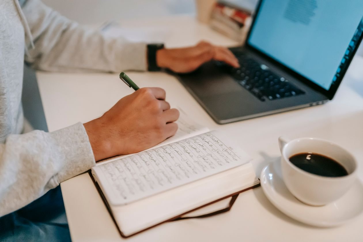 A close-up of a person writing in a notebook while using a laptop, accompanied by a cup of coffee
