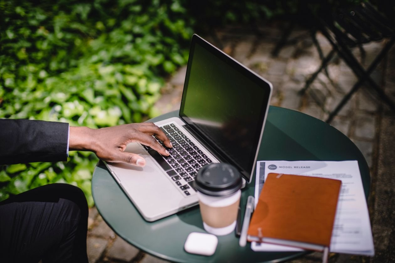 Business professional using a laptop outdoors with a coffee and notebook on a green table
