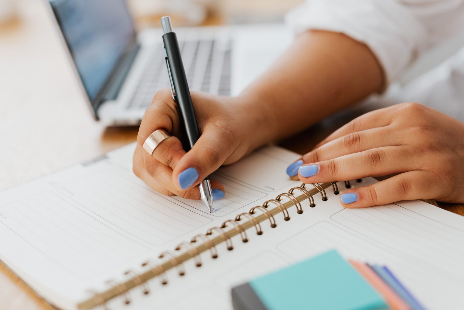 Wanita dengan kuku biru menulis di buku catatan spiral di samping laptop