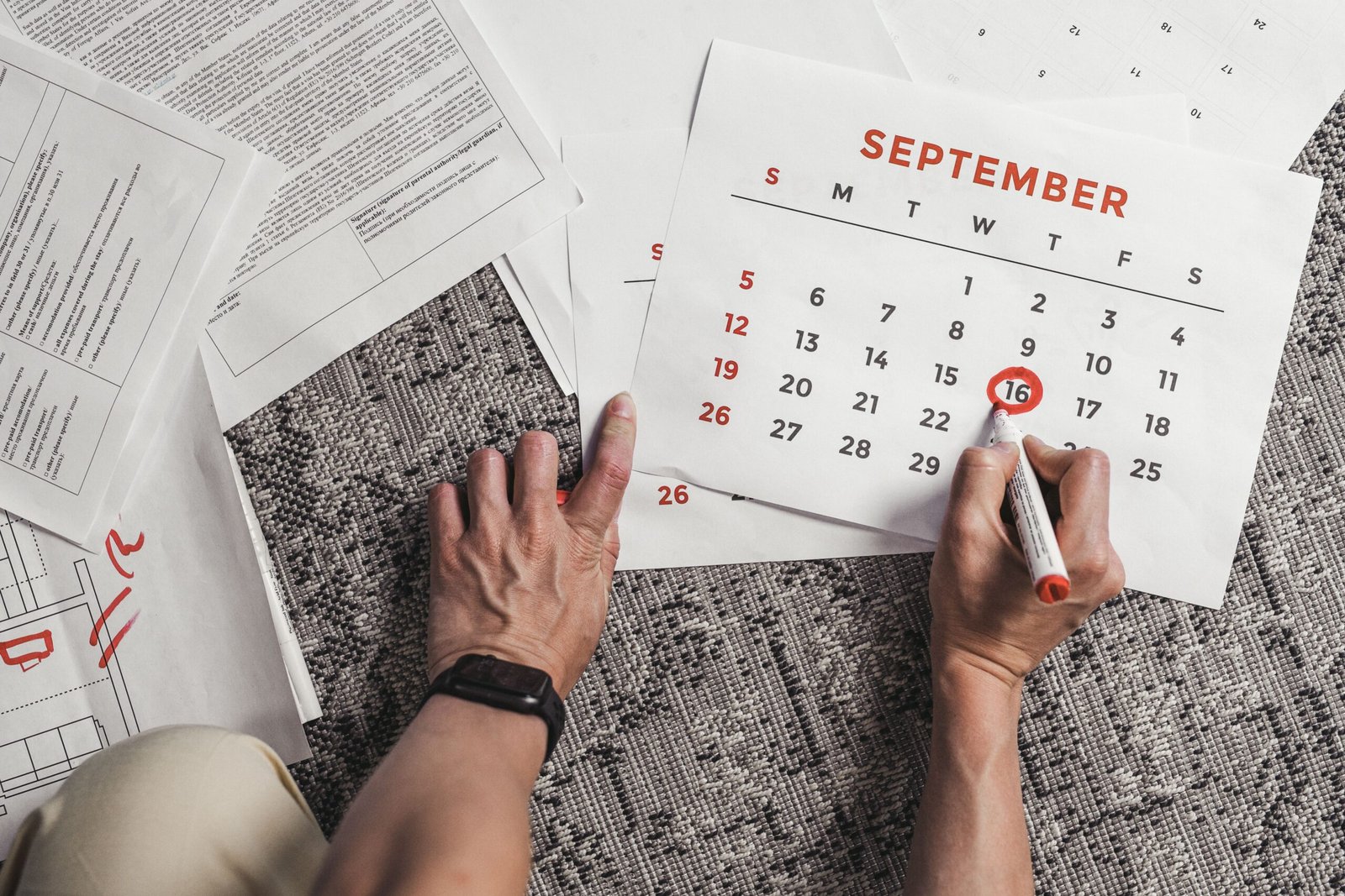 Tangan memegang kalender dengan tanggal September yang dilingkari, dikelilingi oleh dokumen di atas karpet