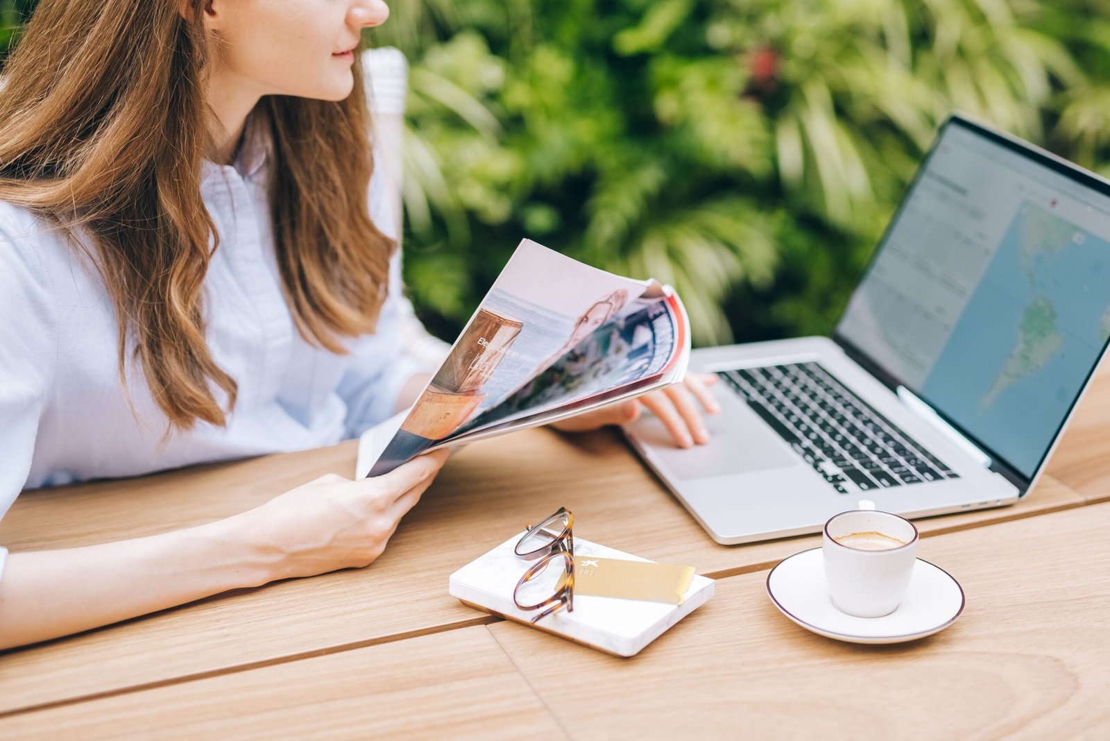 A woman sits outdoors looking at laptop screen, sorrounded by a megazine and coffee