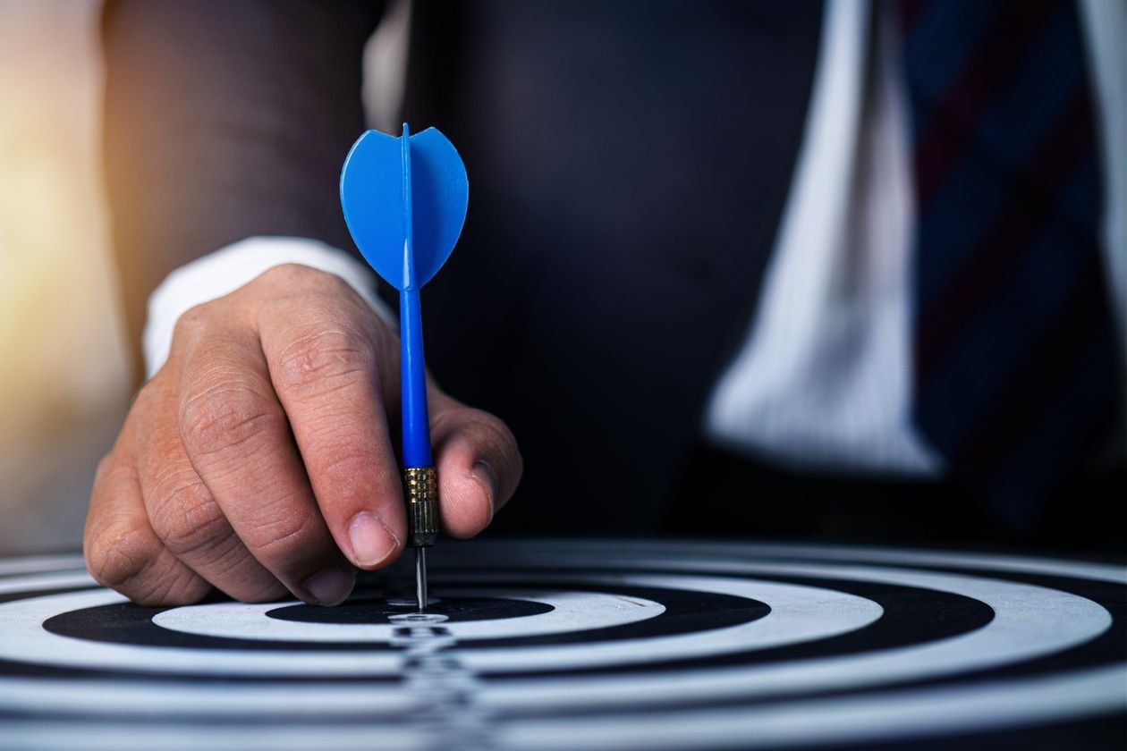 Close-up tangan seorang pria memegang anak panah biru pada bullseye, menyimbolkan target yang dituju.