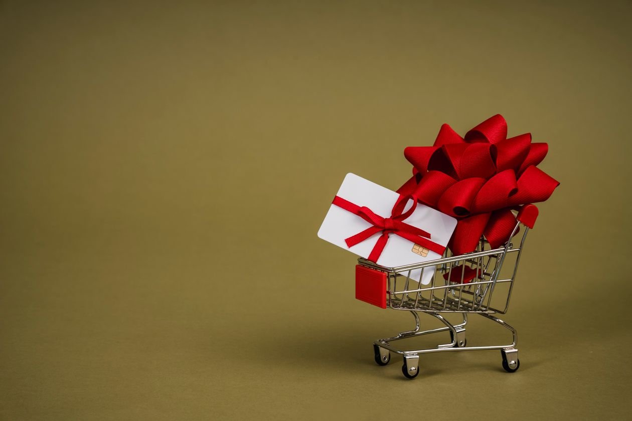 Mini shopping cart containing a gift card adorned with a red bow on a neutral background, ideal for retail concepts