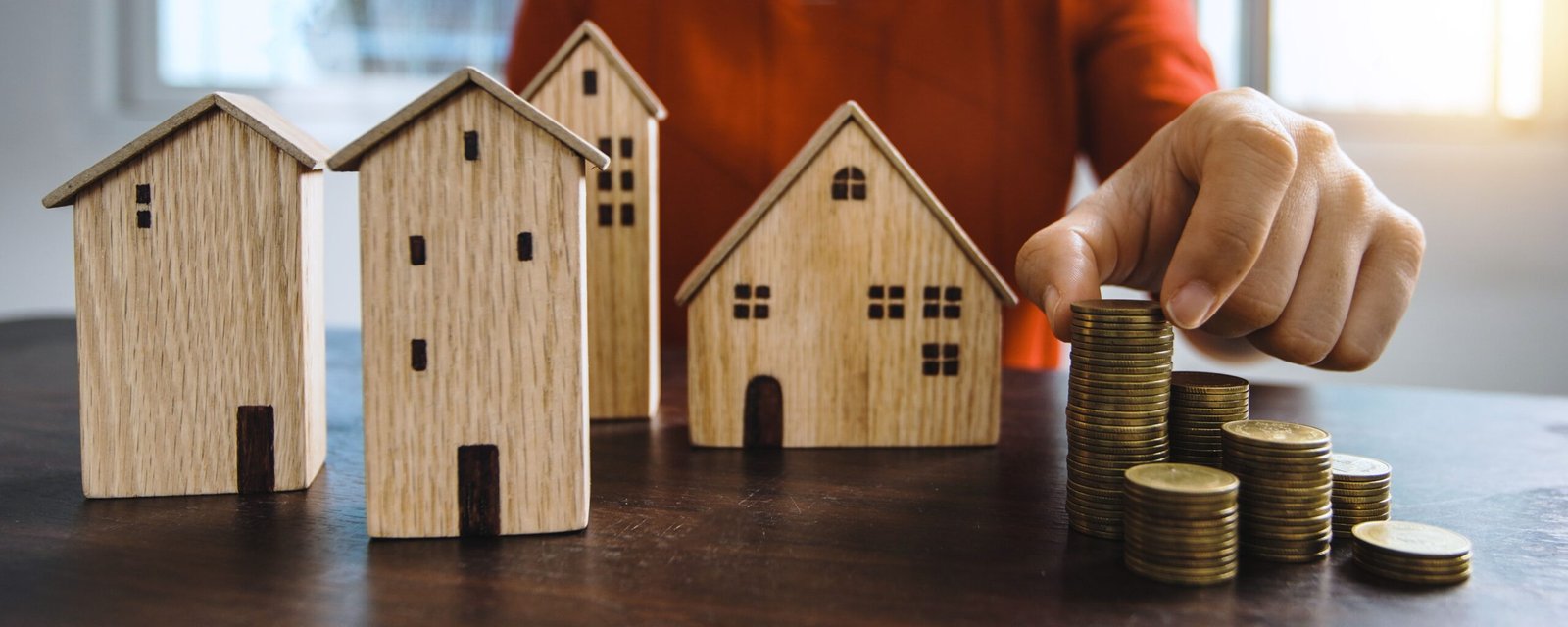 Hand stacking coins next to wooden house models, illustrating budget planning for a property business
