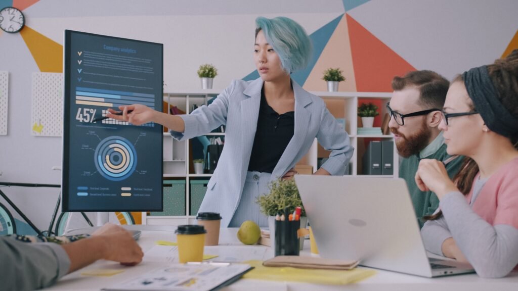 Presentasi analitik bisnis di depan tim, menunjuk grafik persentase di layar monitor dalam rapat kantor