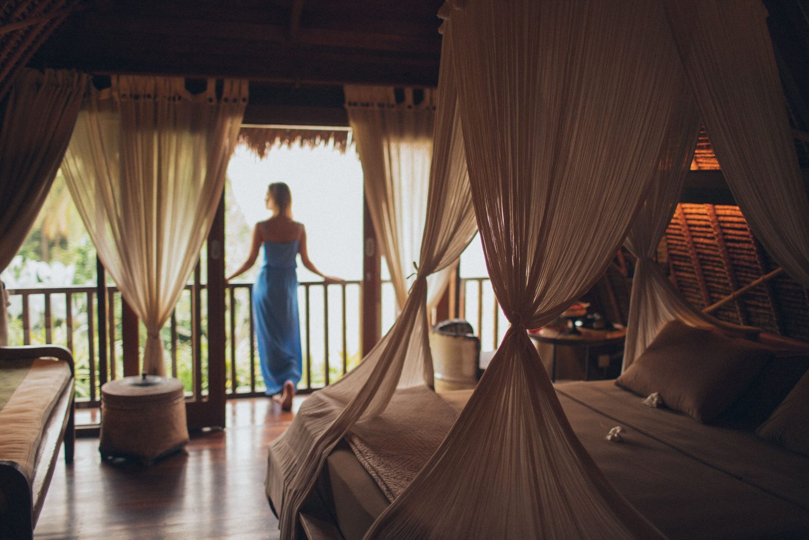Woman standing on the balcony of a tropical villa bedroom with a canopy bed and thatched roof