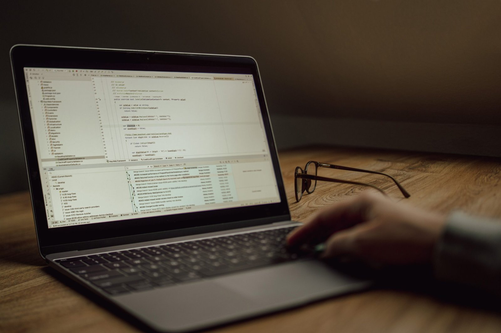 Orang yang mengerjakan kode pemrograman di laptop di dalam ruangan. Kacamata di atas meja